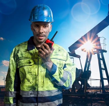 Oil field worker in high-visibility gear using a walkie-talkie near a pump jack at sunset.