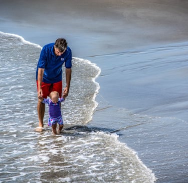 Photo of man holding up baby as they walk through water at the shore
