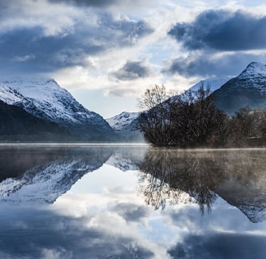 Snowy mountains and clouds reflecting in a calm, misty lake during a winter sunrise.
