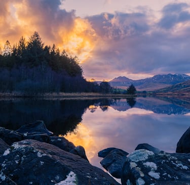 Scenic sunset over a calm lake with mountain reflections and rocky shoreline in the wilderness.