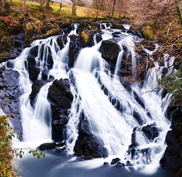 Long exposure photography of a scenic cascading waterfall over dark rocks in a lush autumn forest.