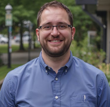 A smiling man with glasses and a beard wearing a blue button-down shirt outdoors.