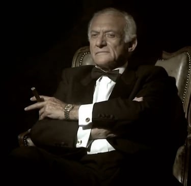 Senolis su švarku, cigaru sėdintis ant kėdės ir žvelgiantis pro šalį