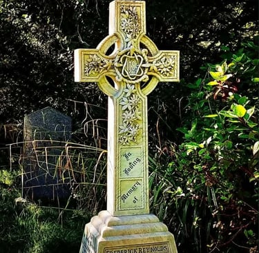 Ivory coloured celtic cross gravestone with ornate flower carving. The background is dense foliage.