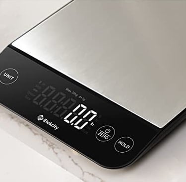 Stainless steel digital kitchen scale with a black display panel showing a zero measurement on a lig