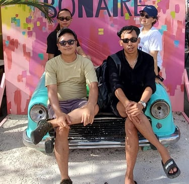 Four people posing on a vintage turquoise car in front of a colorful Cuba Compagnie Bonaire mural.