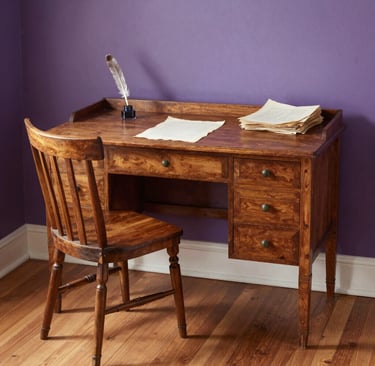 Antique wooden writing desk and chair set against a purple wall with a quill pen and parchment paper.