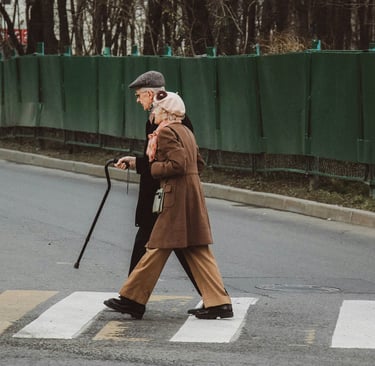 An elderly couple in stylish coats walks across a city street using a pedestrian crosswalk