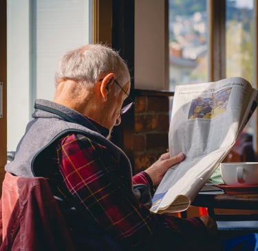 An elderly man in a plaid shirt reading a newspaper at home