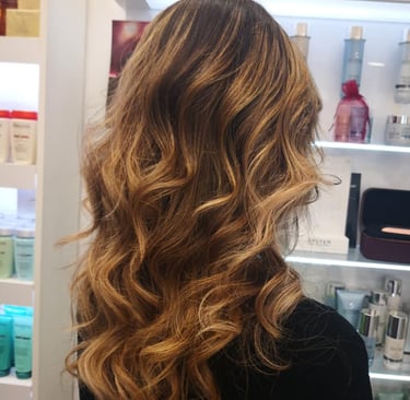 Back view of a woman with long wavy brunette hair and honey blonde balayage highlights in a hair salon.