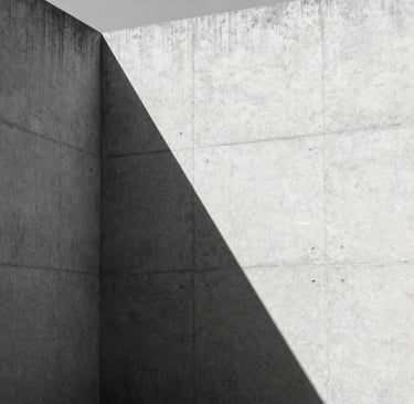 A high-contrast black and white architectural photograph of a minimalist concrete structure in a North American setting. Sharp geometric shadows fall across a light gray textured wall, emphasizing clean lines and sophisticated architectural forms. Editorial style.