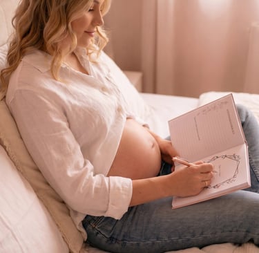 A pregnant woman in a white shirt writes in a maternity journal while sitting on a bed.