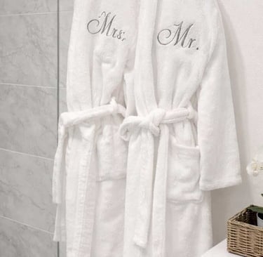 Two white monogrammed robes hang on a bathroom door next to a small orchid and a spa basket