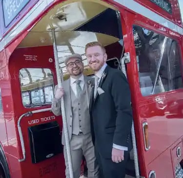 Two grooms in wedding suits smiling at the entrance of a classic red double-decker London bus.