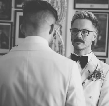 Groom in white tuxedo and bow tie looking at his partner during a wedding ceremony.