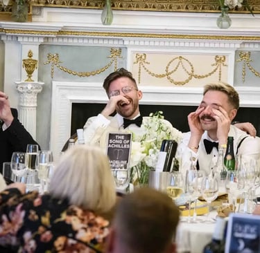 Grooms laughing during wedding speeches at a formal reception dinner table with floral decor.