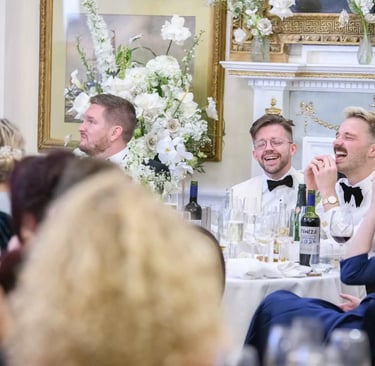 Grooms in white tuxedos laughing at a formal wedding reception dinner table with floral arrangements.
