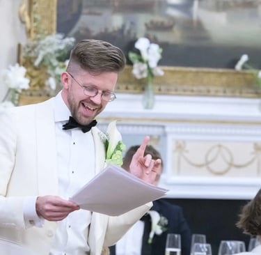 Smiling groom in a white tuxedo giving a wedding speech while holding notes at a reception.