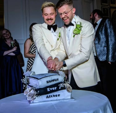 A happy groom couple in white tuxedos cutting their black and white tiered wedding cake.