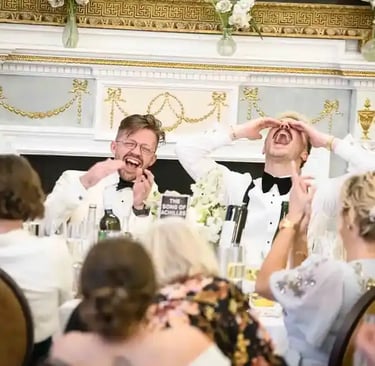 Two grooms in white tuxedos laughing hysterically during wedding reception speeches at a formal head table.