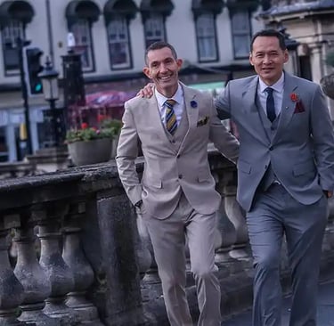 Two stylish men in tailored three-piece suits walking outdoors on a stone bridge.