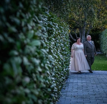 Smiling bride in white lace dress and groom in suit walking on stone path between green hedges.