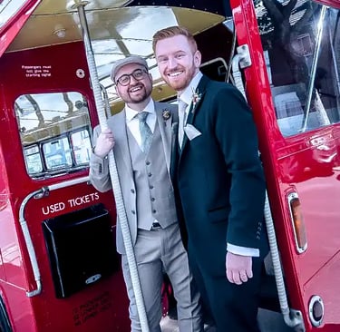 two happy grooms posing on the back platform of a vintage red London bus.
