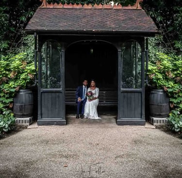 A bride and groom sitting inside a rustic garden gazebo surrounded by lush green foliage.