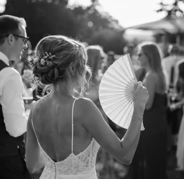 A bride in a lace wedding dress uses a handheld folding fan during an outdoor summer reception.