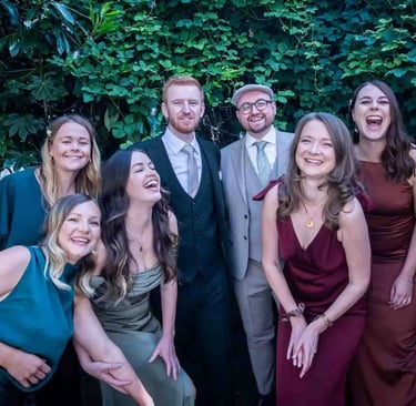 A group of happy wedding guests in formal attire laughing together in a garden setting.