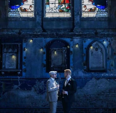 Groom and guest talk inside a rustic wedding venue with stained glass windows and stone walls.