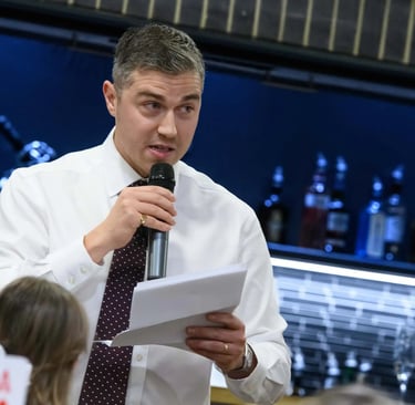 A man in a white shirt and tie gives a formal speech with a microphone and notes at an event.