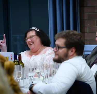 Smiling bride in white lace dress and tiara laughing at a wedding reception dinner table.