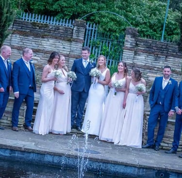 A happy wedding party in blue suits and white bridesmaid dresses posing by a garden fountain.