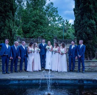 A wedding party in blue suits and pink bridesmaid dresses posing by an outdoor stone fountain.