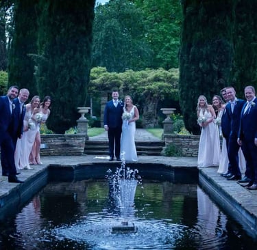 Bride and groom with bridal party posing around a garden fountain at a wedding venue.
