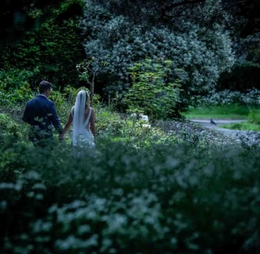 A bride and groom holding hands while walking through a lush green garden at twilight.