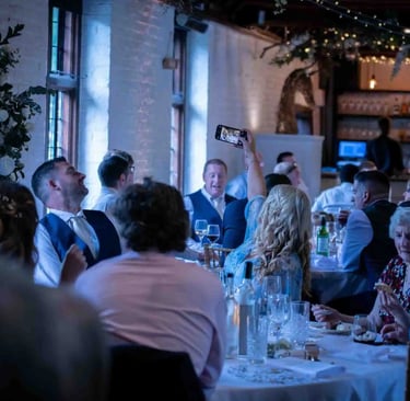 Wedding guests smiling and taking photos with smartphones at a rustic indoor reception dinner.