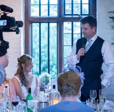 Groom giving a wedding speech at the head table being filmed by a professional videographer.