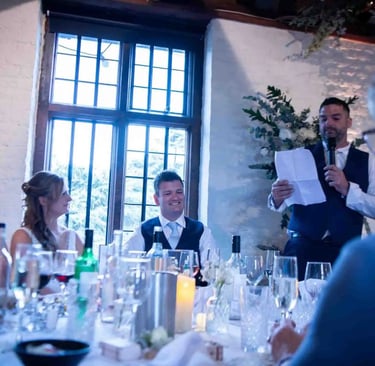 A best man gives a wedding speech at a reception dinner with the smiling bride and groom.