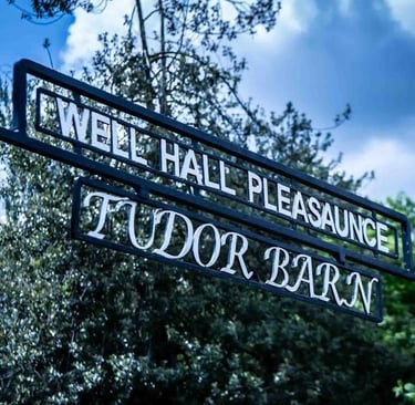 Black wrought iron sign for Well Hall Pleasaunce and Tudor Barn against a blue sky with trees.