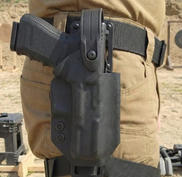 Black kydex tactical holster with a handgun secured on a shooter's belt at an outdoor gun range.