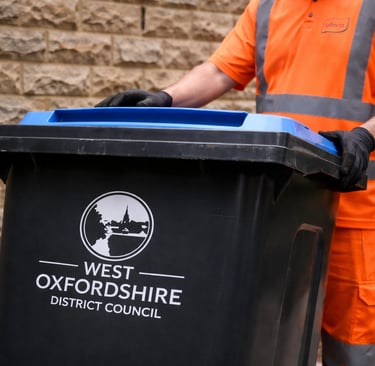 A waste collection worker in hi-vis orange moving a West Oxfordshire District Council recycling bin.