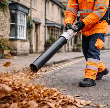 A worker in high-visibility orange gear uses a leaf blower to clear autumn leaves on a street.