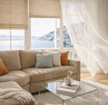 Living room with a beige sectional sofa, Roman shades, and sheer curtains overlooking a coastal ocean view.