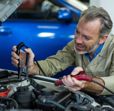 Professional mechanic using jumper cables to jump start a car battery under an open hood.