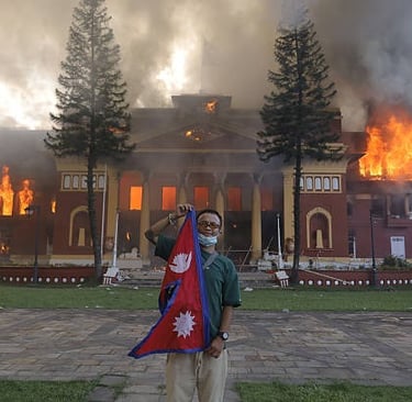 A man holds a Nepal flag in front of a large historic building engulfed in flames and thick smoke.