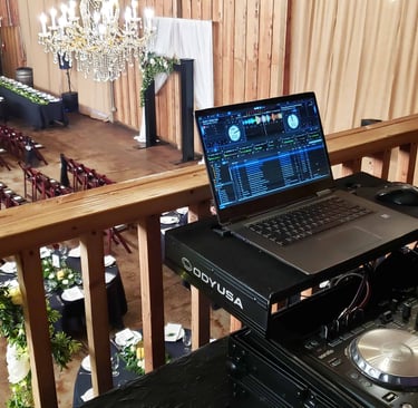 A DJ booth setup on the balcony of a venue overlooking a wedding ceremony space.