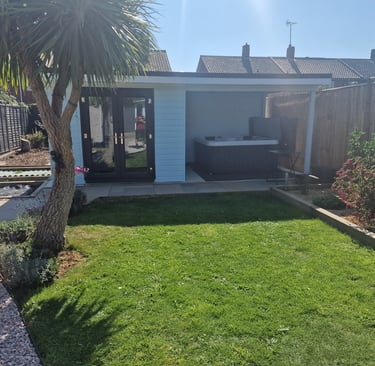 Modern garden cabin with a luxury hot tub and glass doors next to a green lawn and palm tree.