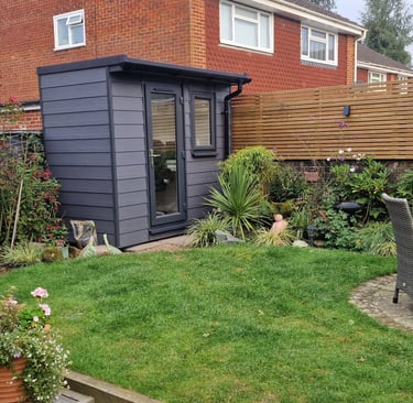 Modern grey garden office pod situated in a landscaped backyard with lawn and patio furniture.